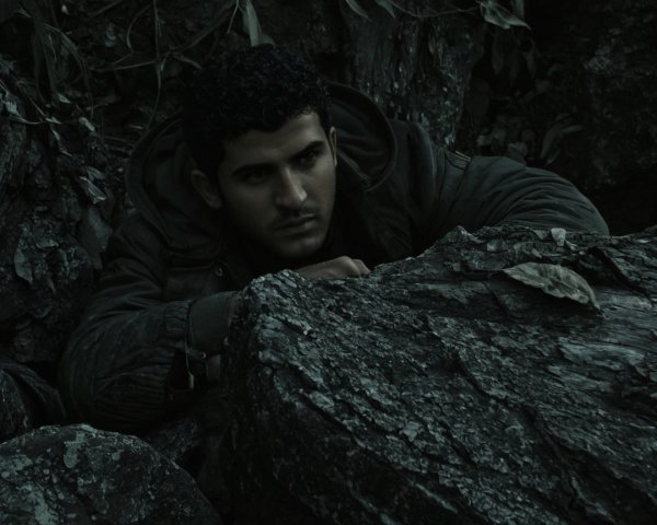 Young man crouched behind rock in shadowy surroundings