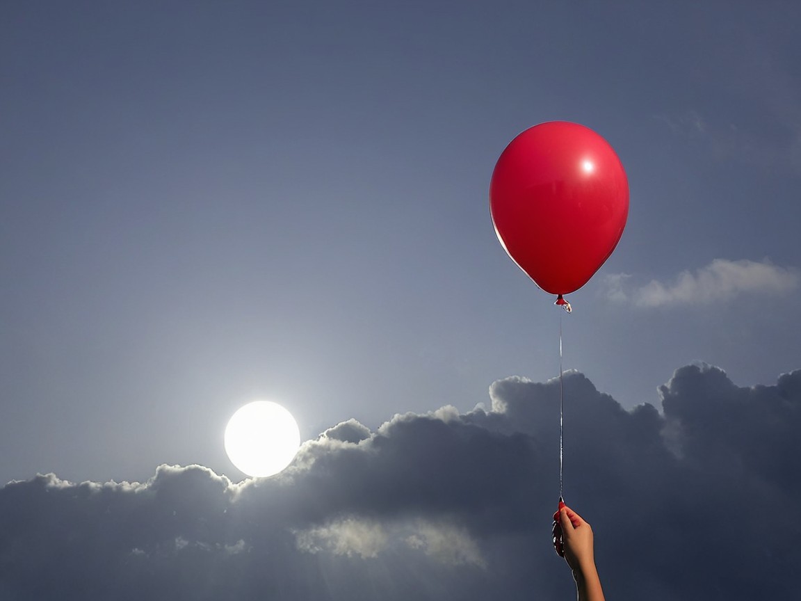 Hand Holding Red Balloon Against Dramatic Sky