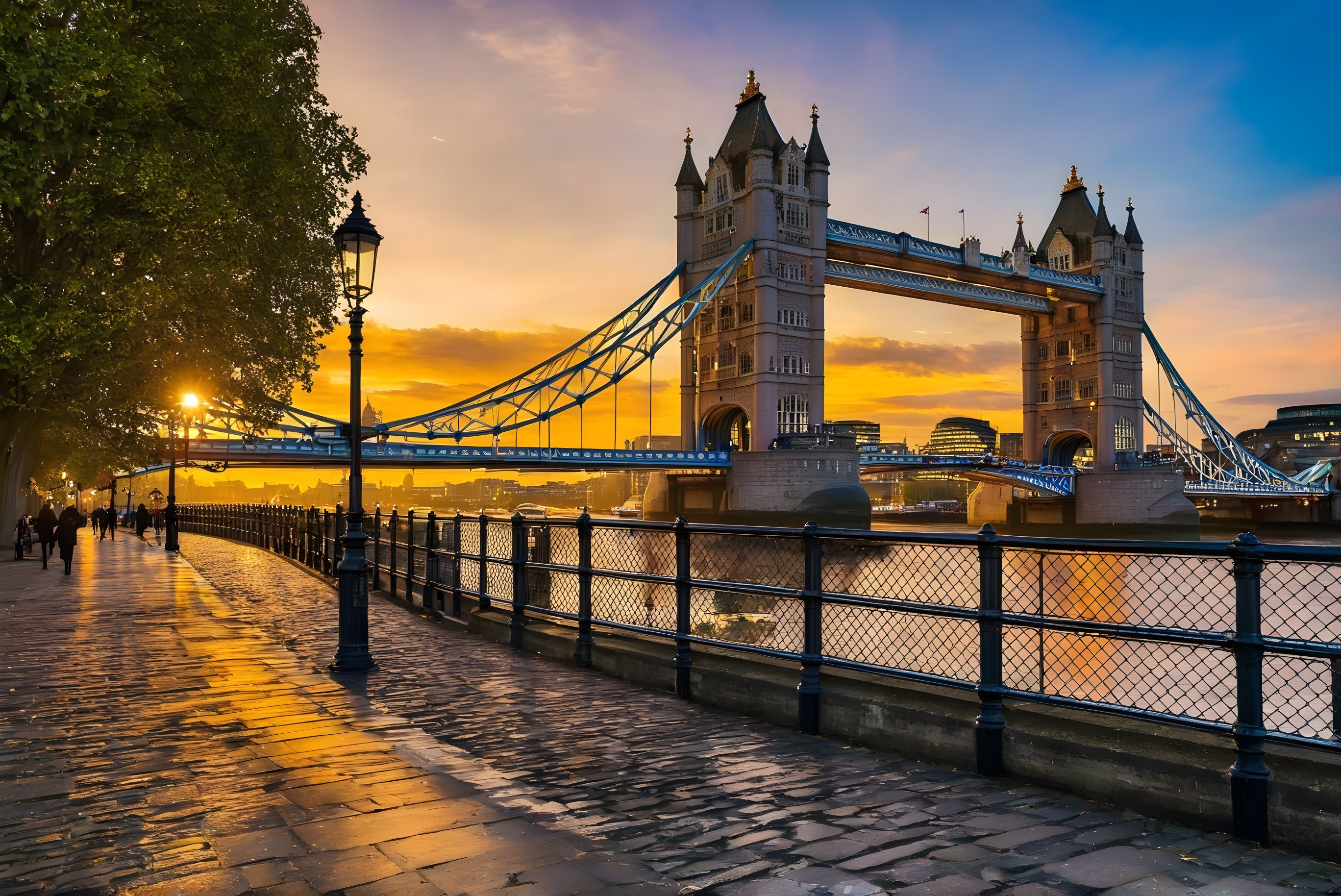 Sunset view of Tower Bridge with glowing sun, lit street lamps, and people by Thames River.